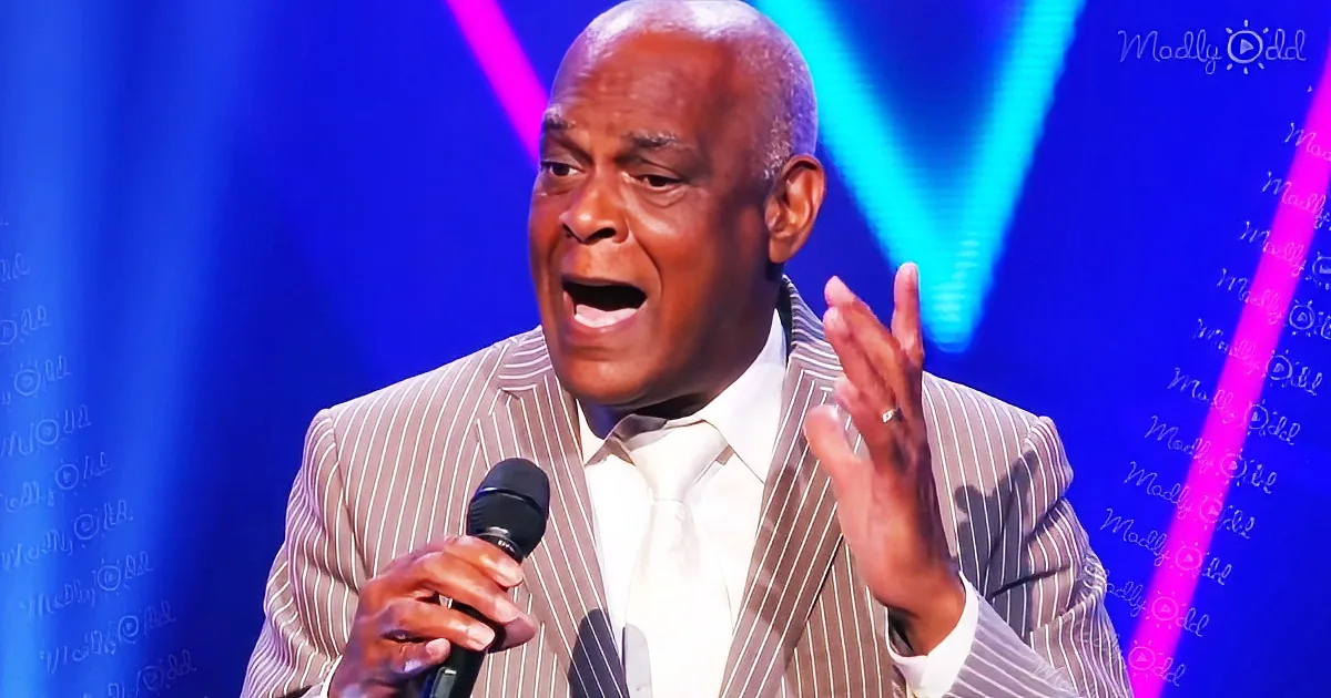 A man in a striped suit holds a microphone, gesturing with his right hand, against a blue stage backdrop.