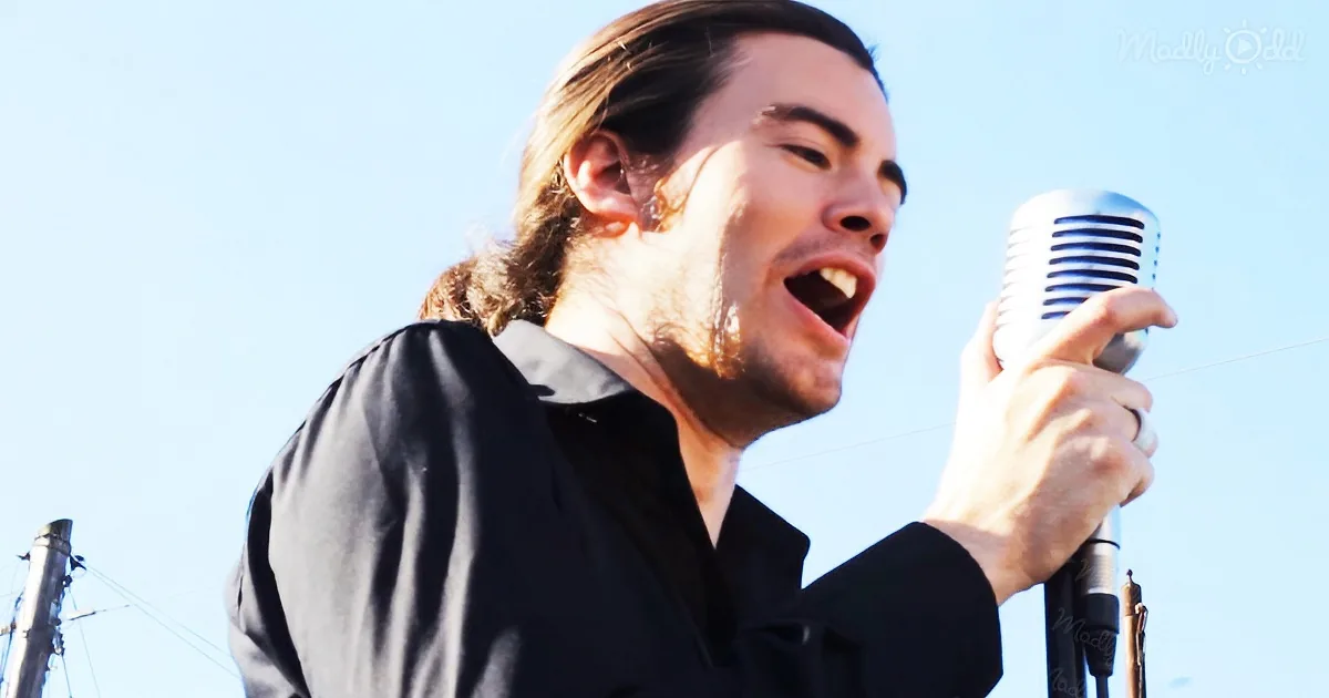 A man with long hair in a ponytail sings passionately into a vintage microphone against a clear blue sky.