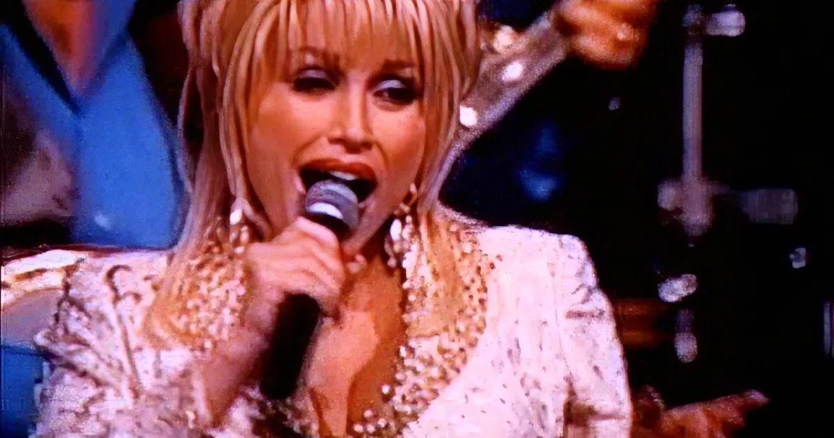 A woman with blonde hair and earrings sings into a microphone, wearing a white lace top, with a guitarist in the background.