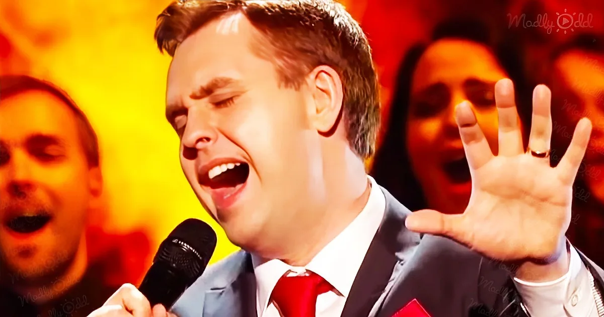 A man in a suit sings passionately into a microphone, raising his hand, with a warm, yellow-lit background.