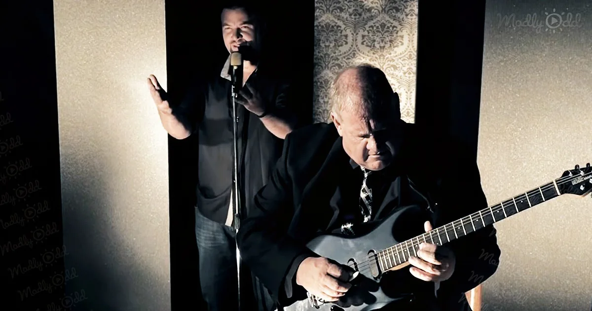 A man in a suit plays guitar while another sings into a microphone.