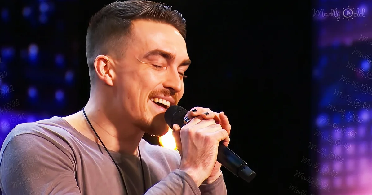 A man with a mustache and short hair holds a microphone, smiling with his eyes closed, wearing a brown shirt.