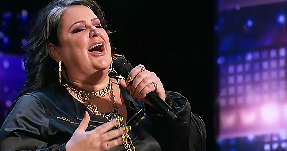 A woman with dark hair and makeup sings passionately into a microphone, adorned with gold jewelry.