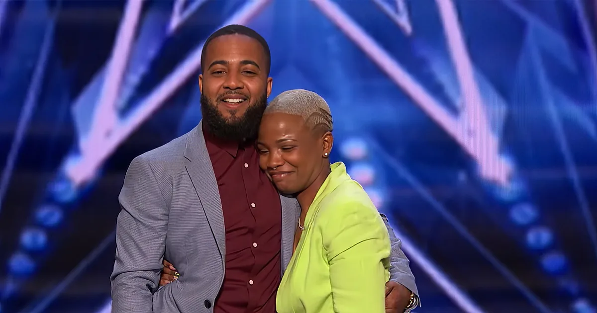 A man and woman smile together on a stage with a colorful background.