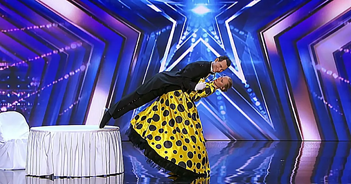A man in a tuxedo balances on a table while a woman in a yellow polka dot dress looks up at him.
