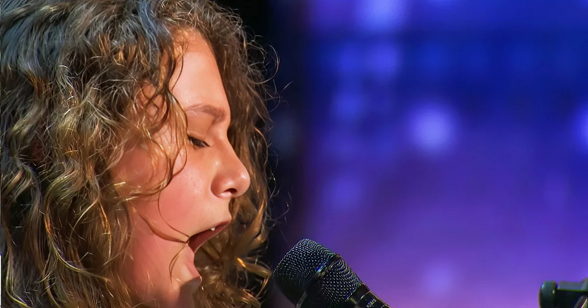 A person with curly hair sings into a microphone, with a blurred blue background.
