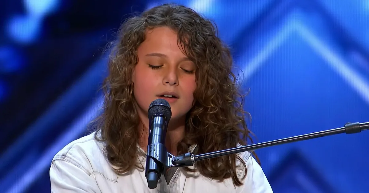 A young person with curly hair sings into a microphone on a stage with blue lighting.