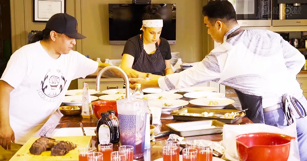 Three chefs in a kitchen preparing food, plates, and drinks on a counter.