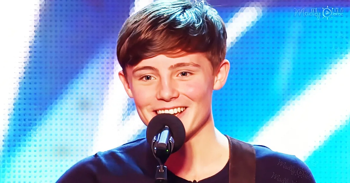 A young person with short brown hair smiles while singing into a microphone on a stage with blue lighting.