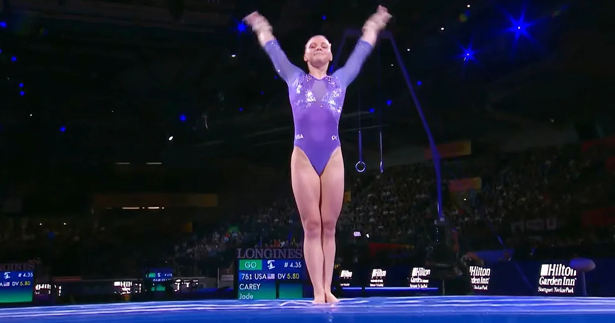 A gymnast in a purple leotard stands on a balance beam with arms raised, surrounded by a dark arena with spectators and...