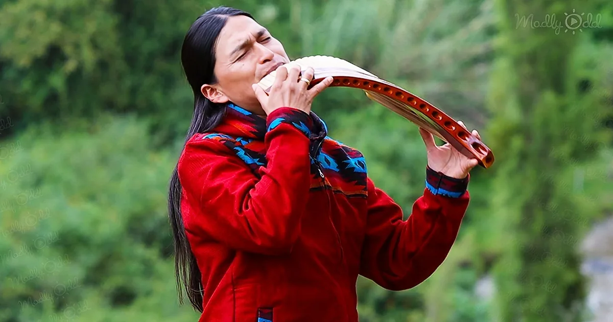 A person in a red jacket plays a wooden instrument outdoors, surrounded by greenery.