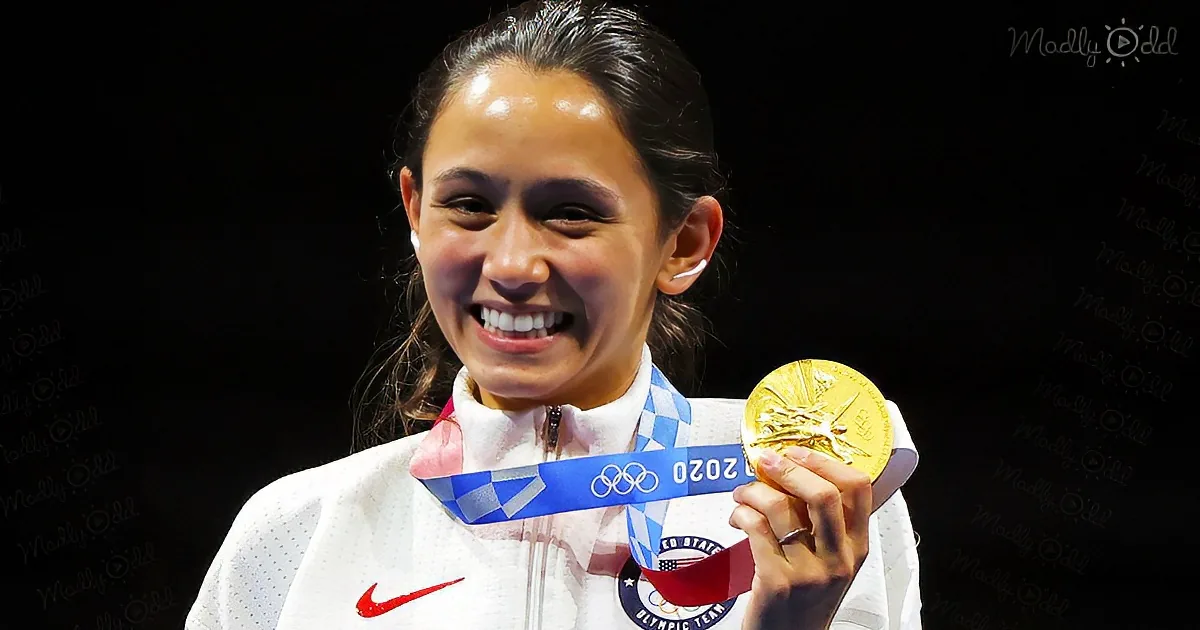 A smiling athlete in a white jacket holds a gold medal with a blue ribbon.