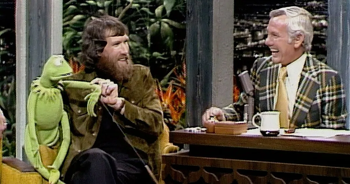 A man in a green jacket is holding a frog puppet, while another man in a plaid shirt laughs nearby.
