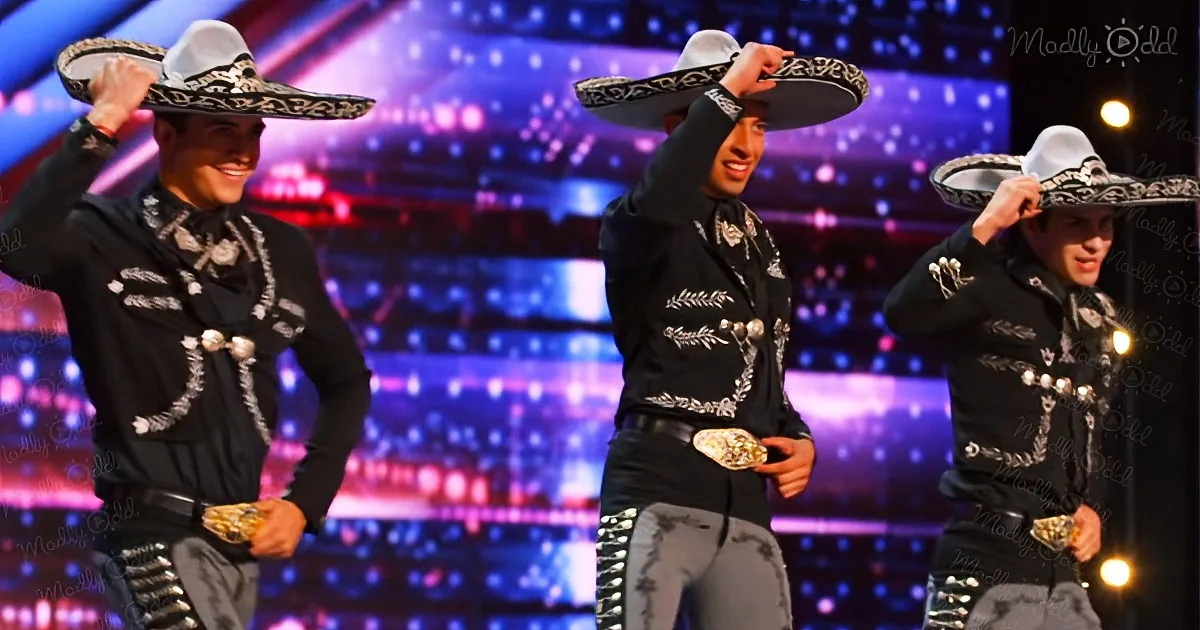 Three men in traditional Mexican attire, sombreros, and silver belts, performing on a brightly lit stage.