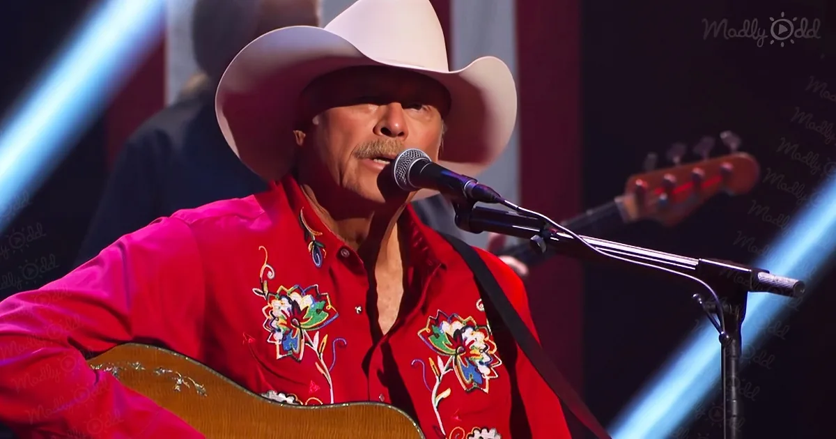 A man in a red shirt and white cowboy hat sings into a microphone while playing a guitar.