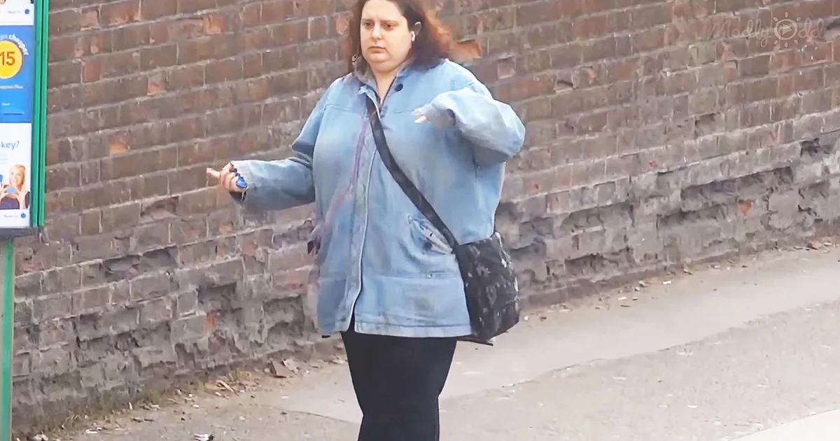 A woman in a blue jacket and black pants walks on a sidewalk, carrying a black bag and holding something in her hand.