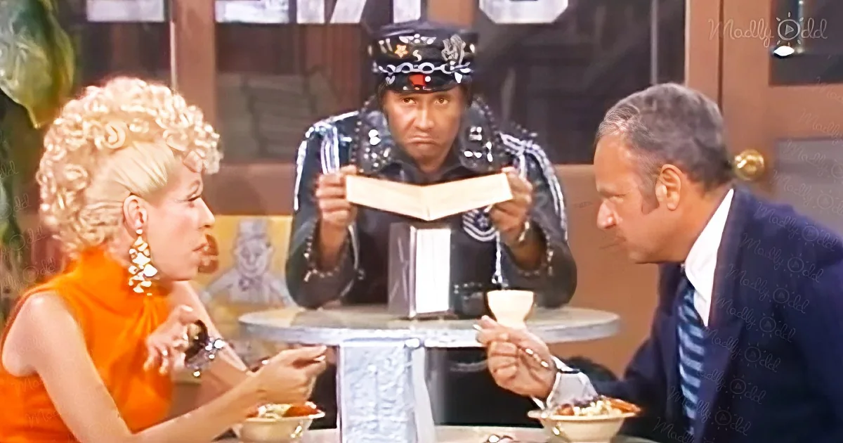 A man in a black outfit and hat holds a menu, while a woman in an orange dress and a man in a suit sit at a table.