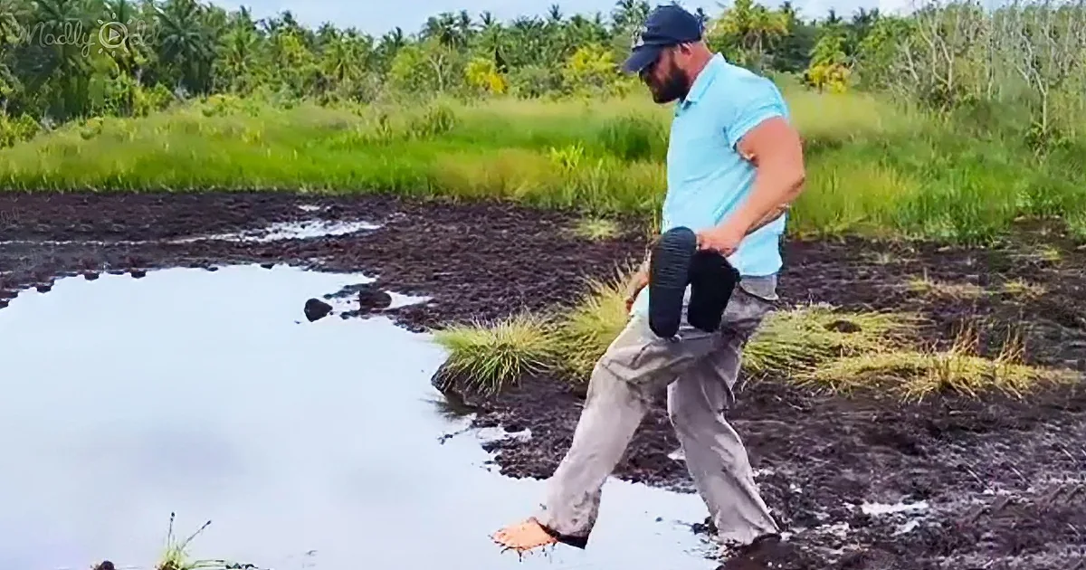 A man walks barefoot through a muddy area, holding a shoe, with a small body of water and lush greenery in the background.