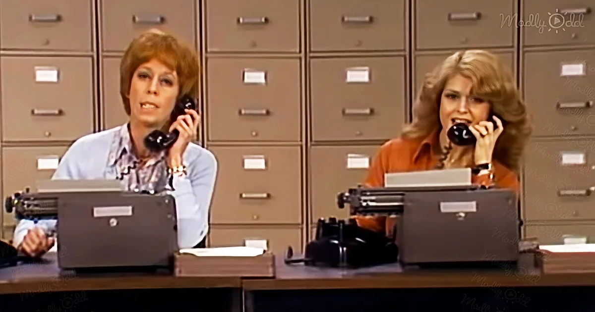 Two women are seated at desks with typewriters, one in a blue shirt and the other in orange, both talking on telephones.