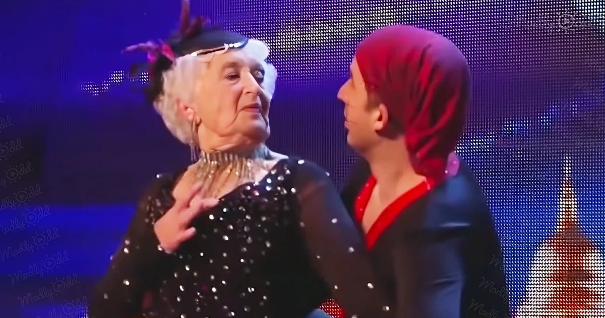 An elderly woman in a black sequined dress dances with a younger partner in a red and black outfit on a stage with a...