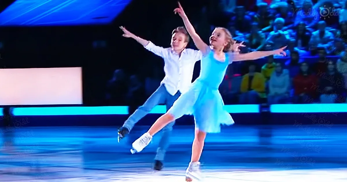 A young girl in a blue dress and a boy in a white shirt skate together on ice, arms outstretched.