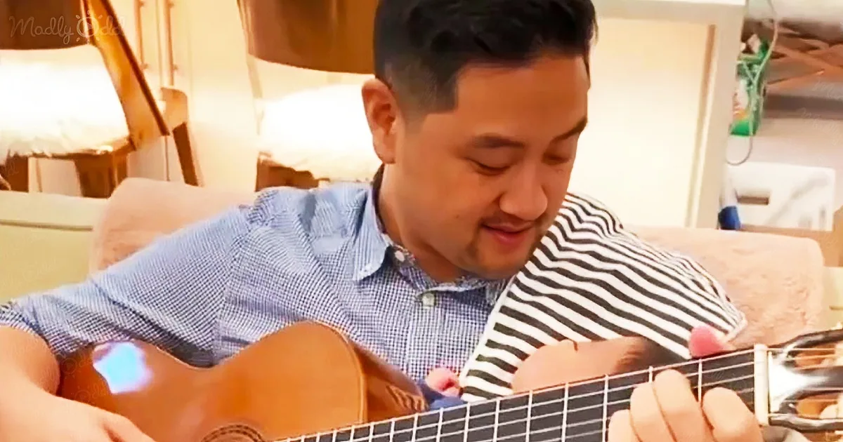 A man in a blue shirt plays an acoustic guitar while a baby in a striped outfit rests on his lap.