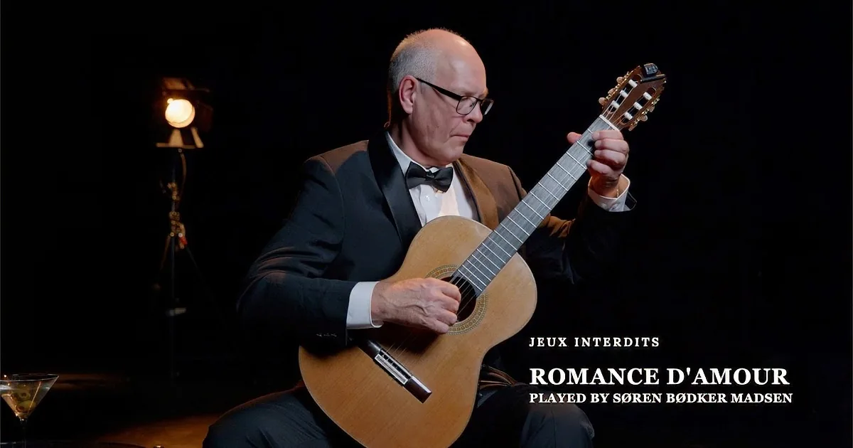A man in a tuxedo plays a classical guitar on stage with a martini glass nearby.