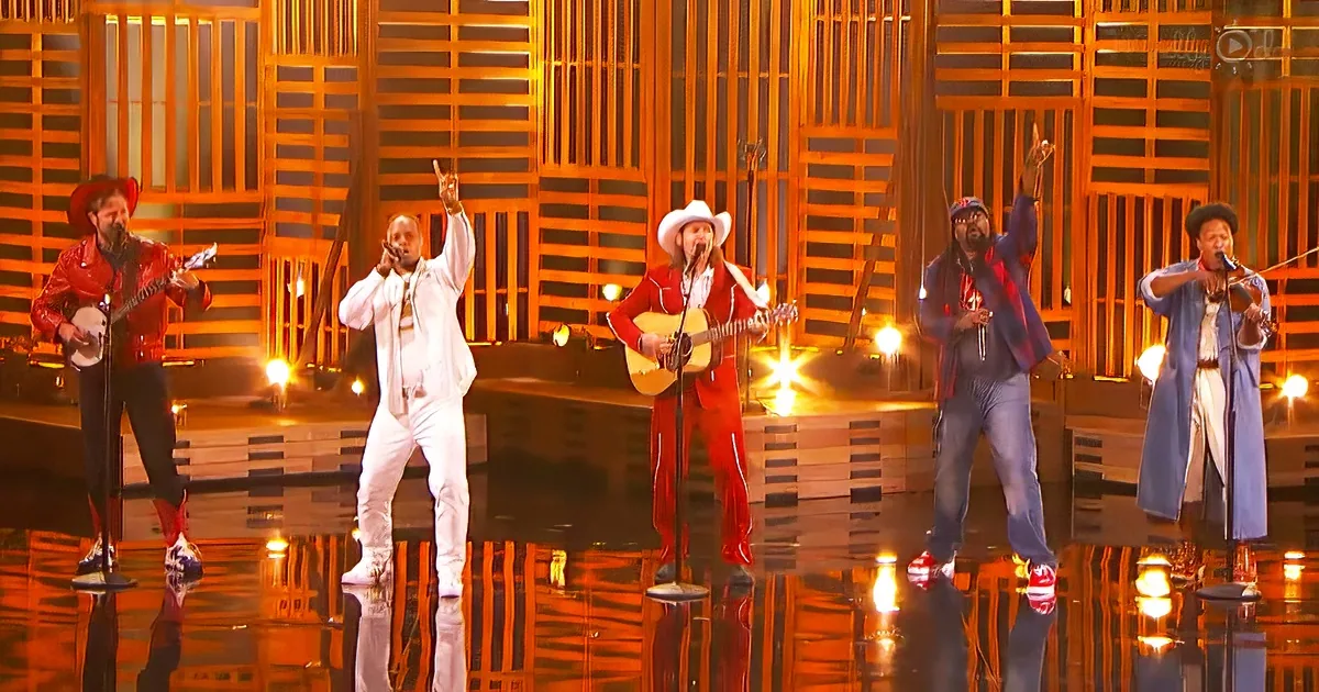 A group of performers in vibrant outfits, including a cowboy hat and red jacket, are singing and playing instruments on a...