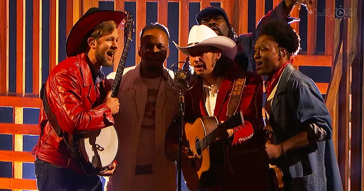 A man in a red leather jacket and cowboy hat plays a banjo, singing into a microphone with a group of musicians.