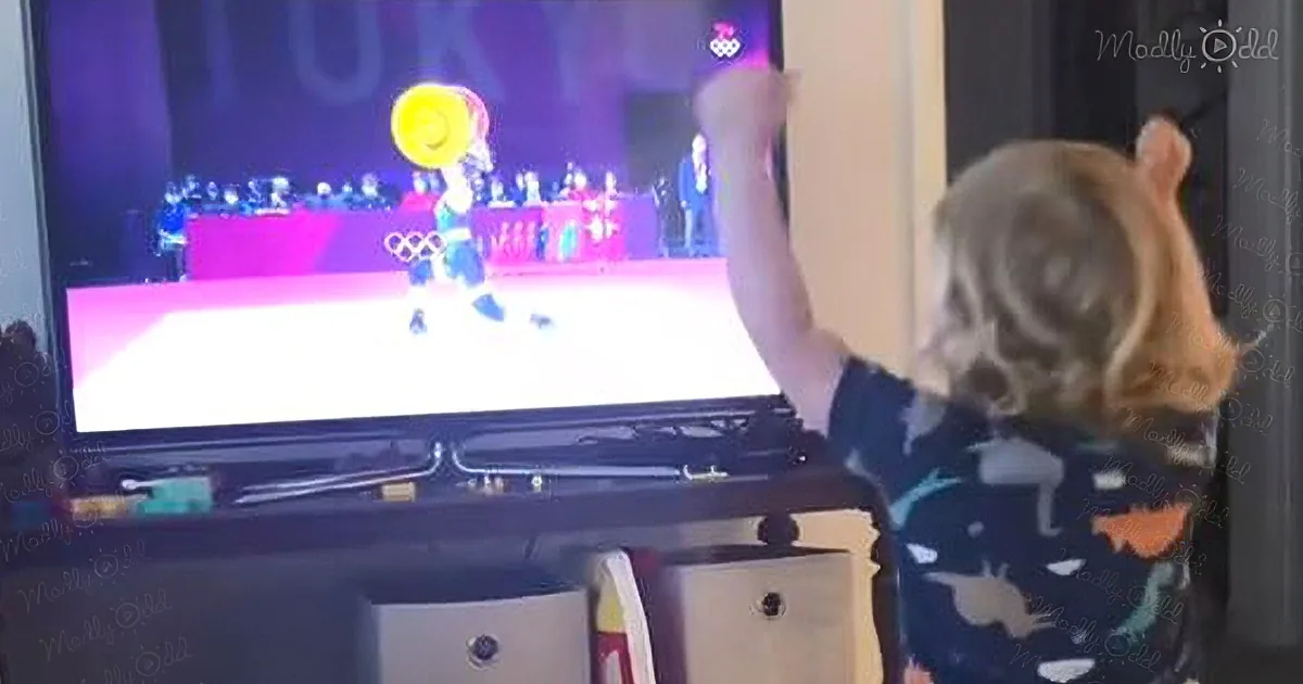 A child mimics a gymnast on TV, holding a yellow disc in front of a large screen.