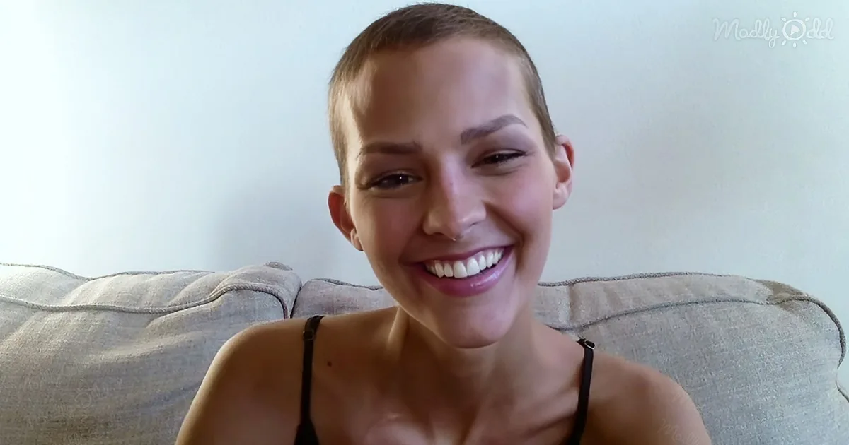 A smiling person with short hair, wearing a black top, sits on a beige couch against a light wall.