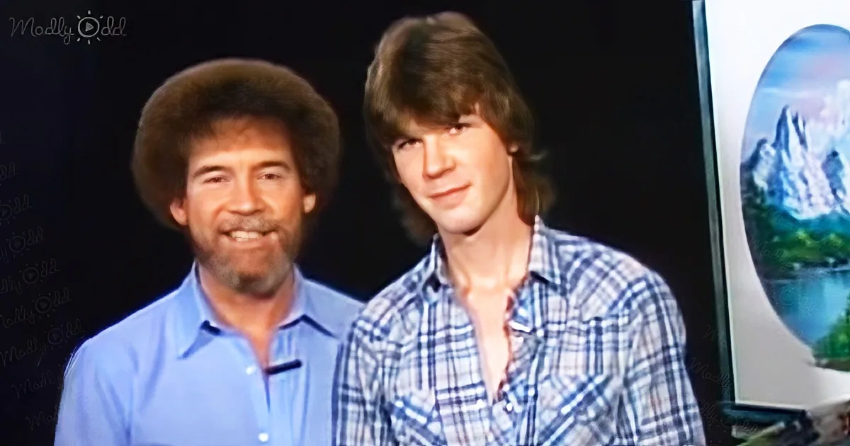 Two men stand together, one in a blue shirt, the other in a plaid shirt, with a painting of a mountain scene in the...