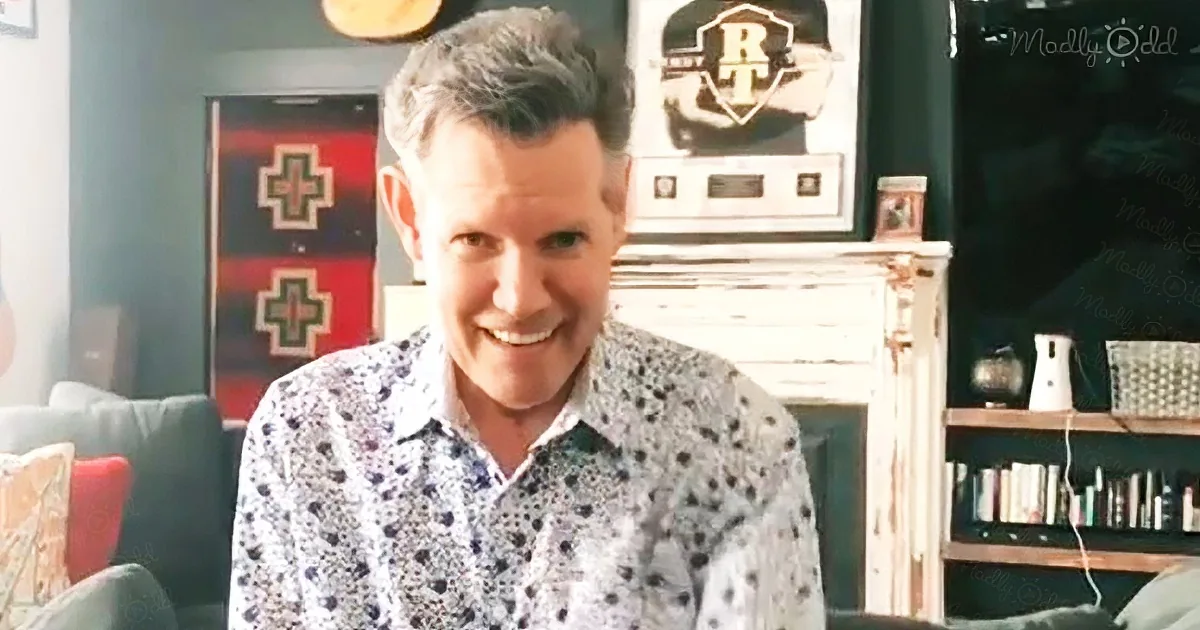 A man in a patterned shirt smiles in a cozy room with a fireplace, books, and a cross-decorated wall.