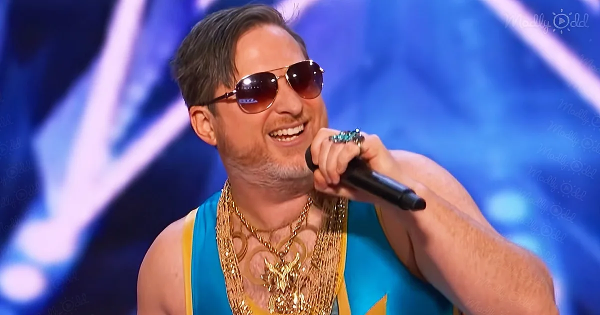 A man in a blue tank top and gold chains smiles while holding a microphone on stage.