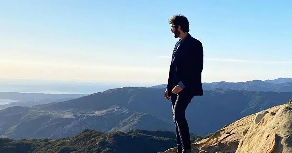 A man in a suit gestures with his hand, standing against a scenic backdrop of ocean and hills.