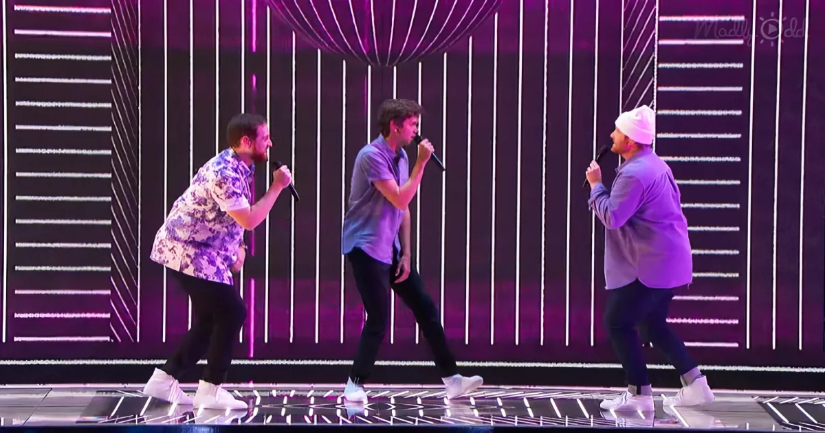 Three men sing on stage with microphones, wearing casual outfits, against a backdrop of vertical white lines and purple...