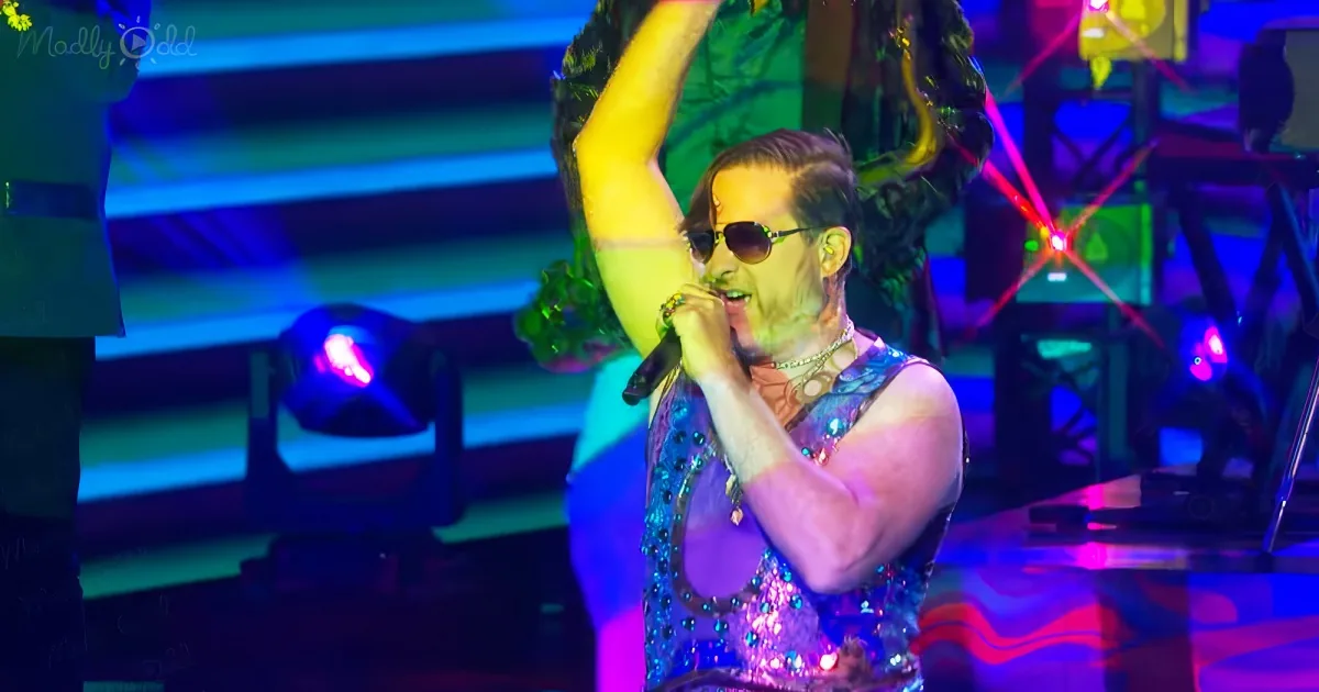 A performer in a colorful outfit sings into a microphone on a stage with vibrant lighting.