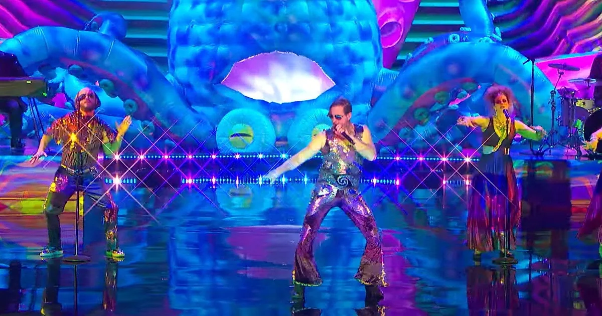 A performer in a shiny outfit sings on stage with colorful lights and inflatable sea creatures in the background.