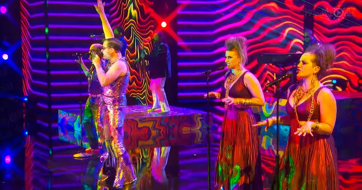 Three performers in colorful, reflective outfits sing on stage with vibrant, abstract background.