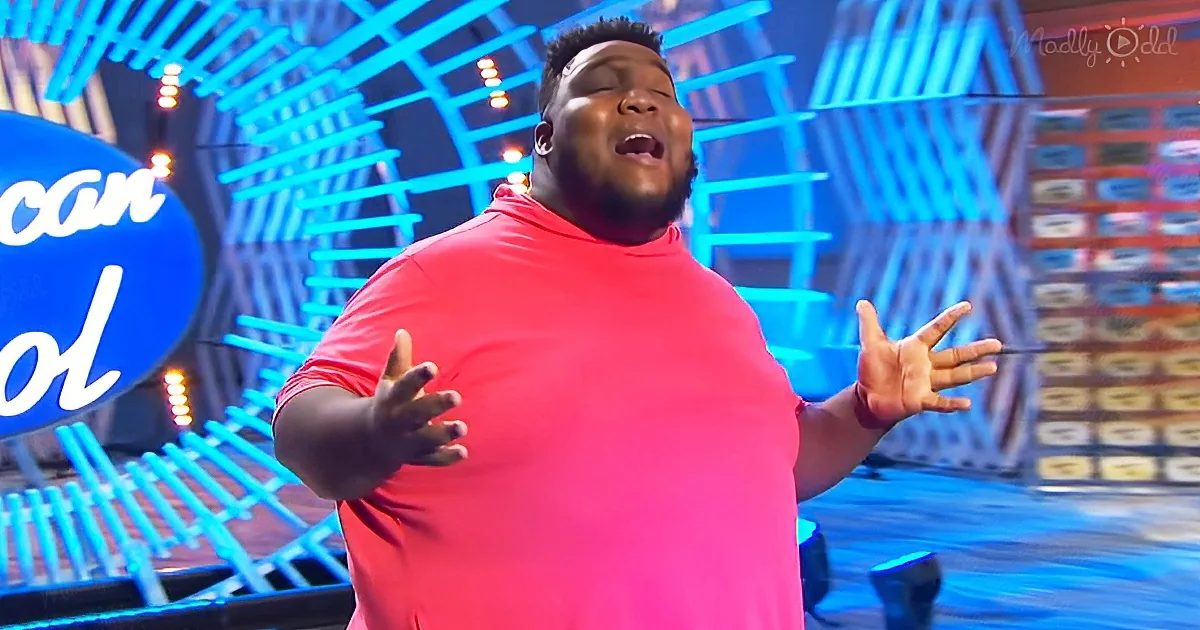 A man in a pink shirt sings passionately on a stage with blue neon lights and an "American Idol" sign.