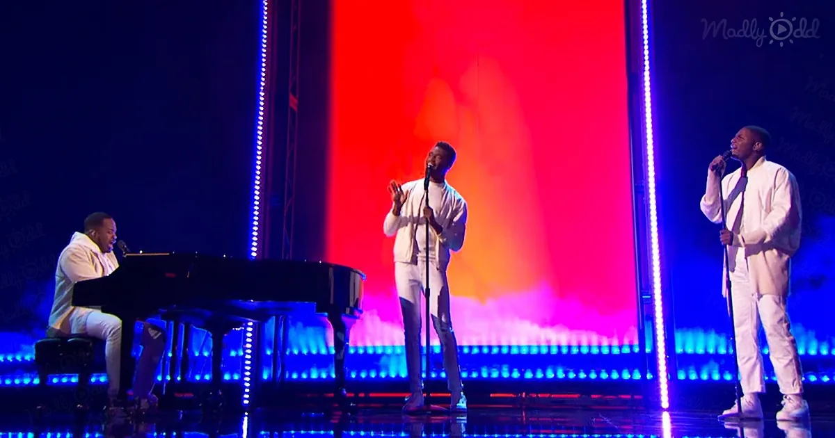A man in white sings passionately on stage with a piano player and another singer, illuminated by vibrant red and blue...