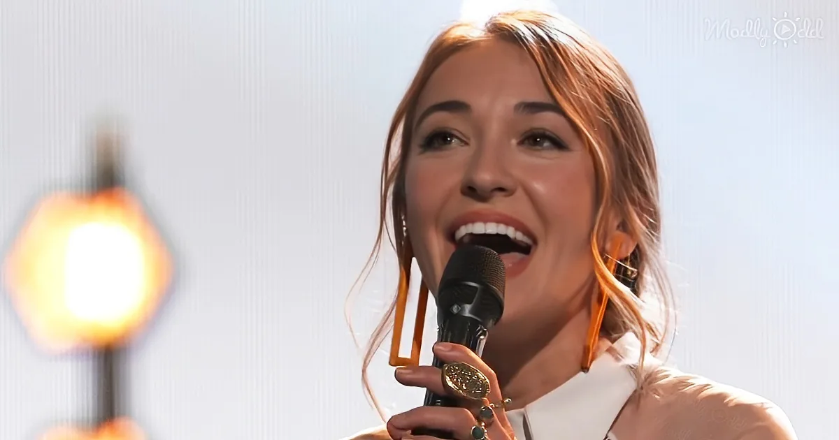 A woman with long hair and earrings sings into a microphone, smiling, with a bright light in the background.