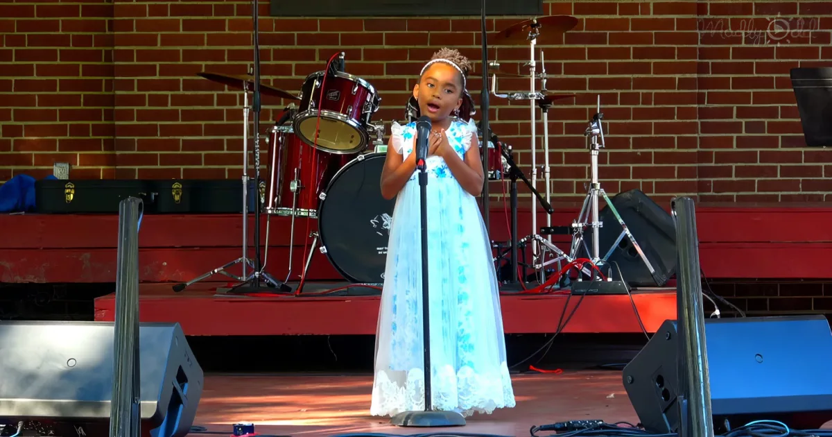 A young girl in a white dress sings into a microphone on a stage with a drum set behind her.