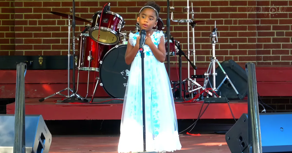 A young girl in a white dress sings into a microphone on stage with a drum set and brick wall behind her.