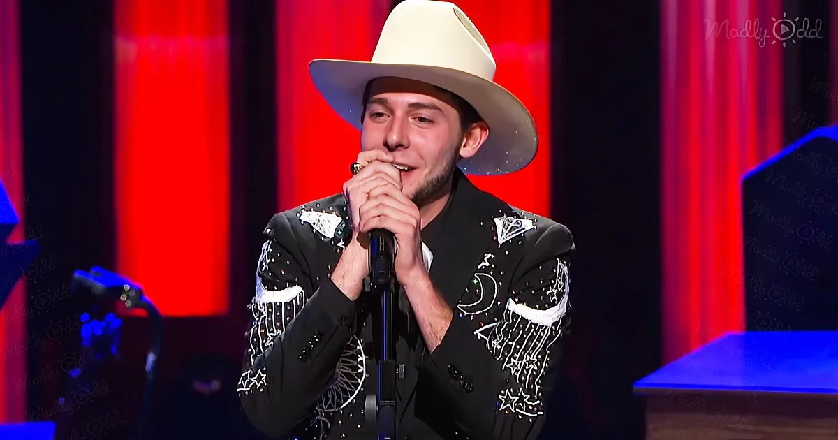 A man in a black suit with white stars and a white hat sings into a microphone on stage with red curtains.