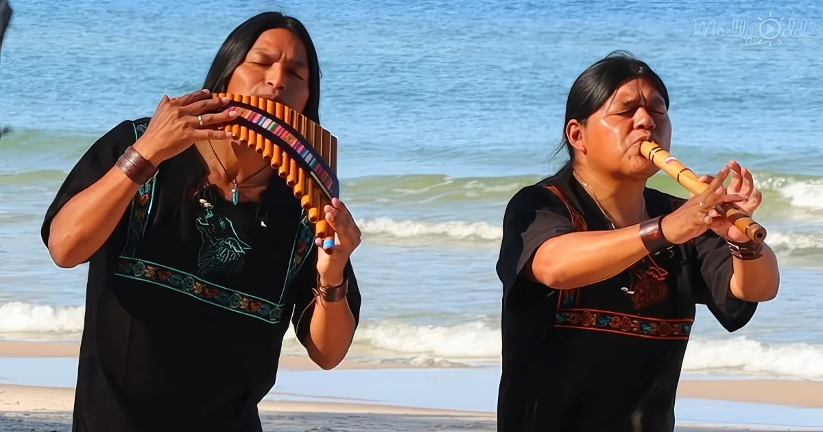 Two individuals play traditional instruments on a beach, with the ocean in the background.