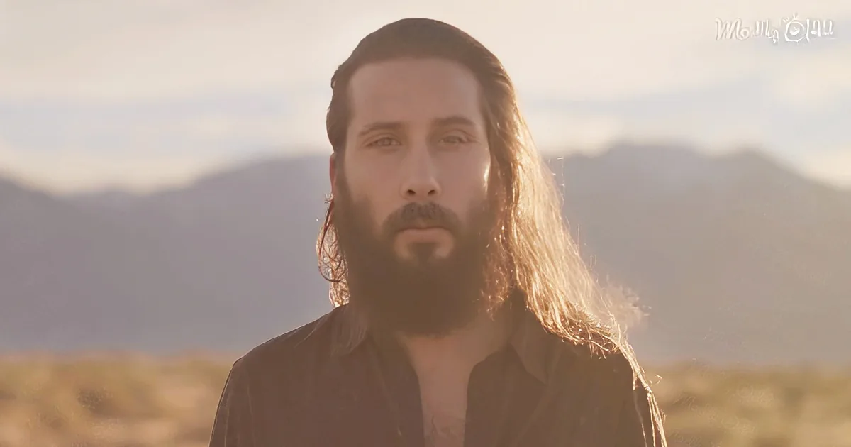 A man with long hair and beard stands in a desert landscape with mountains in the background.