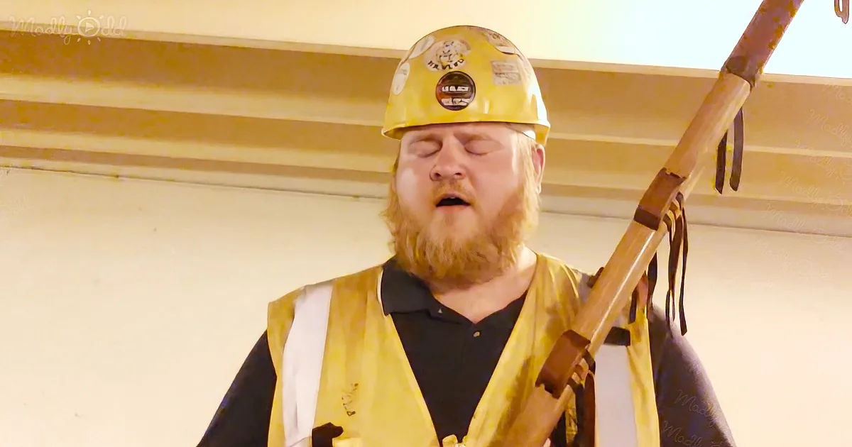 A man in a yellow hard hat and safety vest is holding a long wooden pole, possibly in a construction or industrial setting.