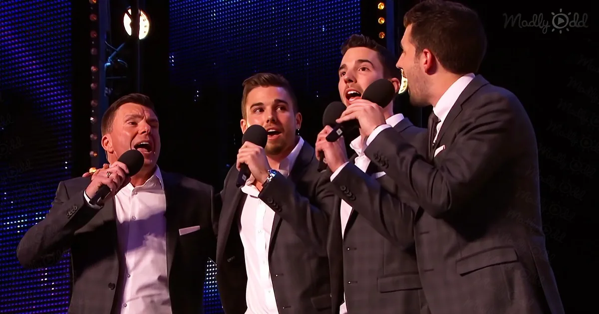 Four men in suits sing into microphones on stage with a LED backdrop.