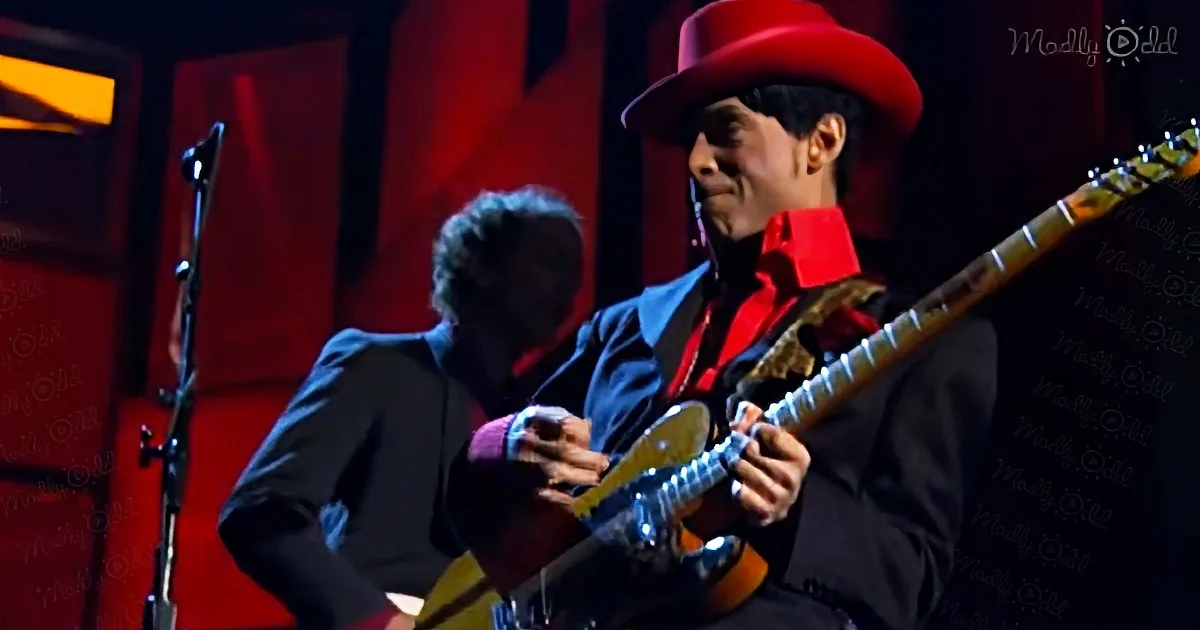 A musician in a red hat and blue suit plays guitar on stage with a red backdrop.